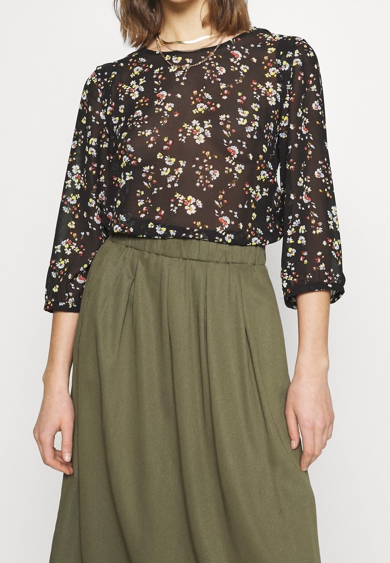 Vrouw draagt een zwarte doorschijnende blouse met bloemenmotief en driekwartmouwen, ingestopt in een olijfgroene rok met hoge taille.