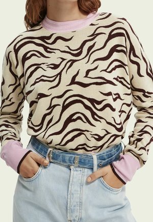 Femme portant un pull beige et marron à rayures zèbre avec des poignets et un col roses, rentrée dans un jean taille haute bleu clair avec une ceinture bleue.