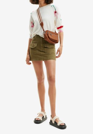 Haut blanc à manches longues avec motifs floraux multicolores, mini-jupe vert olive, sac bandoulière marron et sandales à plateforme noires.