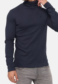 Homme portant un col roulé bleu marine à manches longues et un jean gris clair, ajustant le col d'une main.