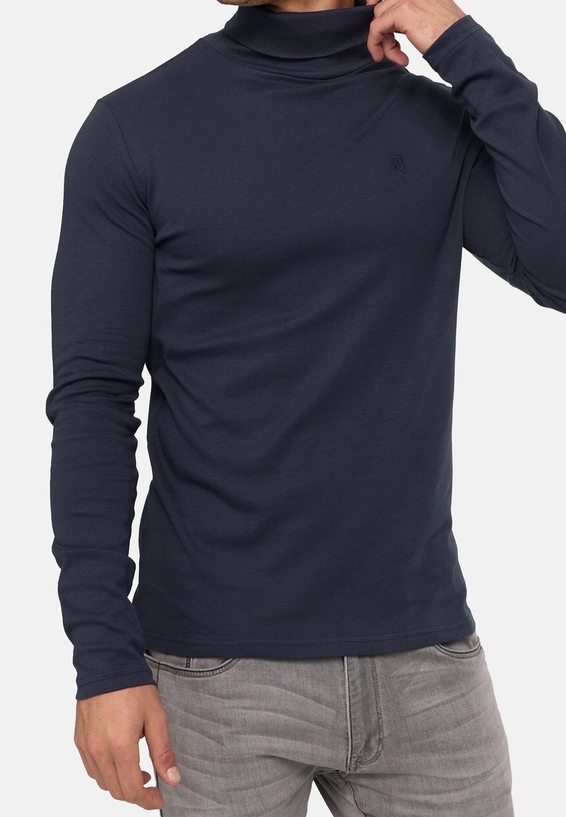 Homme portant un col roulé bleu marine à manches longues et un jean gris clair, ajustant le col d'une main.