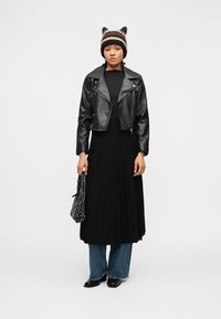 Γυναίκα που φοράει μαύρο δερμάτινο μπουφάν, μαύρη πλισέ φούστα πάνω από μπλε τζιν, μαύρες μπότες, ριγέ πλεκτό καπέλο με αυτιά και κρατά μια τσάντα με καρφιά.