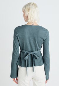 Grön texturerad långärmad topp med rund halsringning, peplumstil och en knuten rosett bak. Kombinerad med ljusa byxor.