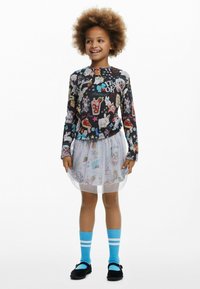 Langärmliges schwarzes Oberteil mit bunten Tier- und Lebensmittelmotiven; weißer Tüllrock; blaue Socken mit weißen Streifen; schwarze Schuhe mit Akzenten.