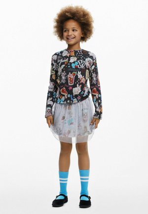 Langärmliges schwarzes Oberteil mit bunten Tier- und Lebensmittelmotiven; weißer Tüllrock; blaue Socken mit weißen Streifen; schwarze Schuhe mit Akzenten.