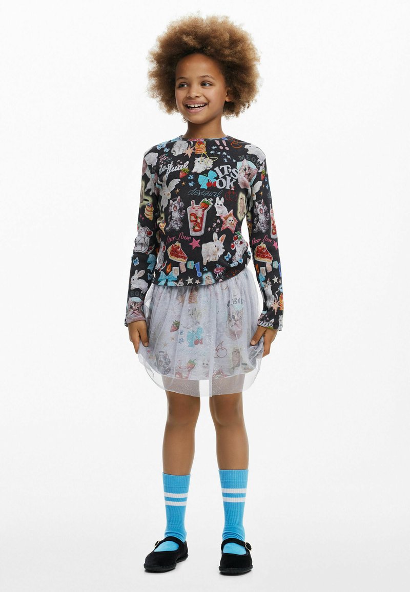 Langärmliges schwarzes Oberteil mit bunten Tier- und Lebensmittelmotiven; weißer Tüllrock; blaue Socken mit weißen Streifen; schwarze Schuhe mit Akzenten.