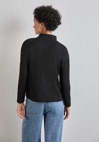 Top lungo a maniche lunghe nero a costine con colletto alto e fessure laterali, abbinato a jeans di denim blu chiaro. Design semplice, senza motivi o accenti.