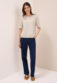 Kvinde med mørkt hår står i en beige kortærmet top, mørkeblå jeans og brune sneakers foran en lysfarvet hjørnevæg.