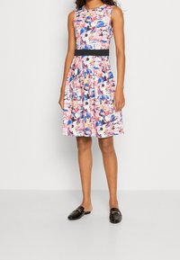 Robe fleurie sans manches; présente des motifs roses, bleus et crème, un corsage ajusté et une ceinture foncée. Associée à des chaussures plates noires.