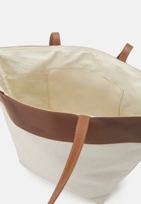 Plátěná taška na rameno s jemným koženým lemováním; má jednoduchý tvar, dlouhé popruhy a kapsu uvnitř podšívky v světlé barvě.