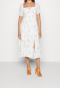 Robe florale blanche avec un col carré, des manches bouffantes et une fente haute sur la cuisse, associée à des sandales à talons oranges.