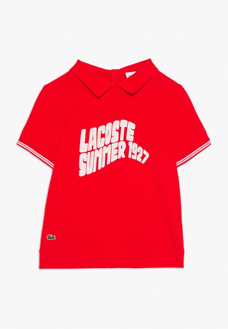 Czerwona koszulka polo z klasycznym kołnierzykiem, z białym napisem "LACOSTE SUMMER 1927". Krótkie rękawy z białymi akcentami; logo Lacoste na dole.