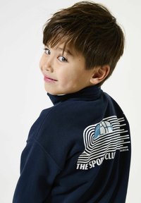Marineblauwe sweatshirts met een hoge kraag, met een graphic op de achterkant die golven, strepen en de tekst "THE SPORT CLUB" toont.