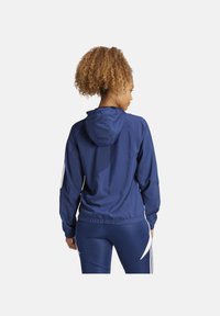 Giacca a zip di colore blu navy con cappuccio, polsini e vita elasticizzati. Presenta strisce bianche decorative sulle maniche e sul retro. Tessuto liscio e leggero.