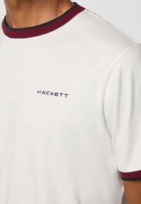 Biela bavlnená tričko s okrúhlym výstrihom a vínovo-modrým ribovaným lemovaním. Na hrudi vyšívané logo "HACKETT" v námorníckej modrej farbe.