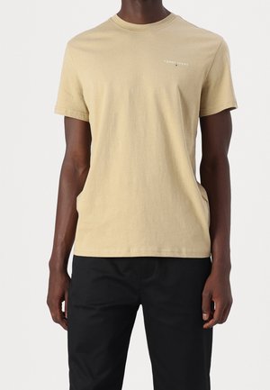 Man draagt een beige Tommy Jeans T-shirt met korte mouwen en zwarte broek, staand tegen een effen lichte achtergrond.