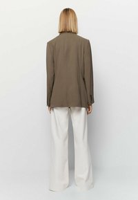 Blazer marrone su misura con spalle strutturate, spacco posteriore singolo e polsini a bottone piccoli, abbinato a pantaloni bianchi a gamba larga.