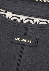 Czarna skórzana torebka z teksturowaną powierzchnią, złotym logo Coccinelle oraz kontrastową, wzorzystą podszewką widoczną przy otwarciu.