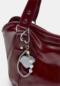 Geantă de mână din piele de un roșu închis, cu un pandantiv în formă de inimă din oglindă argintie și o brățară din mărgeluțe atârnând de curea.