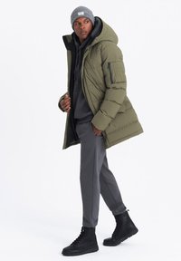 Chaqueta de plumón verde olivo con capucha y patrón acolchado, usada sobre una sudadera con capucha gris oscuro y pantalones a juego, combinada con botas negras de cordones.
