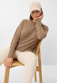 Brauner Langarmpullover aus weichem Material, kombiniert mit hellbeigen Cordhosen und einer hellrosa Baseballkappe. Sitzt auf einem Holzstuhl.