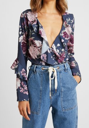 Mujer con una blusa floral de escote en pico profundo y puños con volantes, metida dentro de unos vaqueros azules de talle alto con un cinturón blanco de cordón.