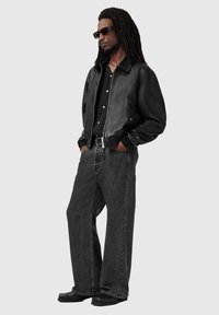 Czarna skórzana kurtka typu bomber na czarnej koszuli zapinanej na guziki, zestawiona z luźnymi ciemnoszarymi dżinsami i czarnymi butami. Model nosi okulary przeciwsłoneczne.