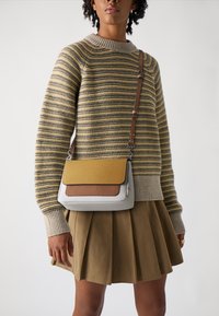 Krusta soma pelēkā ādas ar teksturētu brūnu un sinapsa dzeltenu vāku. Iespējas ar noņemamu brūnu siksnu un strukturētu dizainu. Redzama svītrainā džemperis.