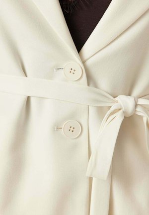 Kremfarvet blazer med to runde knapper og et matchende stofbælte bundet i en knude ved livet over en mørk top.