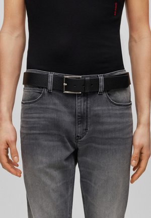 Persoon draagt verweerde grijze spijkerbroek en een zwarte leren riem met een zilveren rechthoekige gesp.