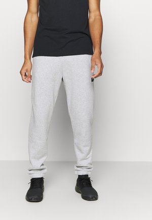 Persoon die lichtgrijze joggingbroek, zwarte sneakers en een zwart T-shirt draagt, staand tegen een effen witte achtergrond.