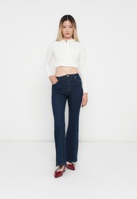 Bijela skraćena veste s rebrastom teksturom, u paru s tamnoplavim trapericama širokih nogavica. Model nosi crvene sjajne cipele; obična bijela pozadina.