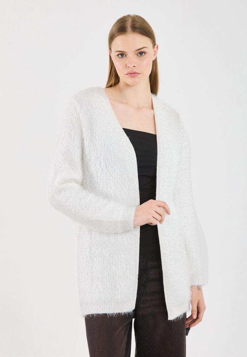 Cardigan alb cu un material texturat și pufos, cu design deschis în față, mâneci lungi și o croială relaxată. Purtați peste un top negru.