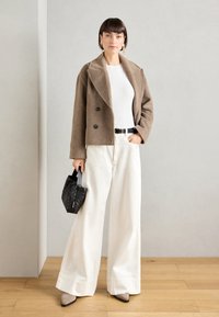 Veste double boutonnage en mélange de laine marron sur un haut blanc, associée à un pantalon large blanc. Sac à main noir texturé et chaussures pointues beiges.