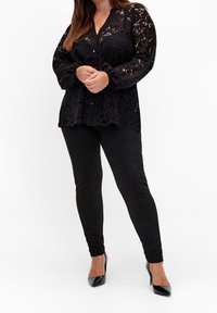 Kvinna iförd svart spetsblus med långa ärmar, svarta åtsittande byxor och svarta lackade högklackade skor, stående mot vit bakgrund.