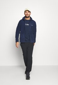 Navy fleece jacket with a high collar, worn over a dark blue t-shirt featuring white text. Black pants and dark shoes complete the outfit.