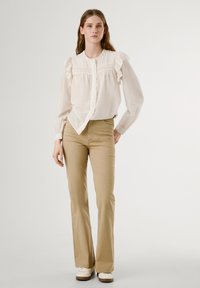 Weiße Bluse mit Rüsche an den Schultern und Knöpfen, kombiniert mit hellbeigen, ausgestellten Hosen und weißen Turnschuhen. Glattes Material und lässige Passform.