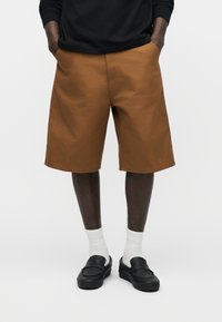 Άντρας που φορά καφέ σορτς μέχρι το γόνατο, λευκές κάλτσες και μαύρα παπούτσια χωρίς κορδόνια, στέκεται με τα χέρια στις τσέπες μπροστά σε μονόχρωμο φόντο.