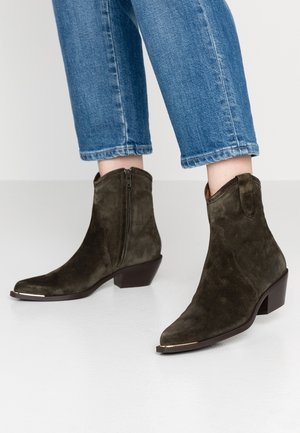 Personne portant des bottines en daim brun foncé avec des fermetures éclair latérales et de petits talons carrés, associées à un jean bleu court.