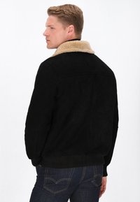 Schwarze Wildlederjacke mit einem beigefarbenen Lammfellkragen, gerippten Bündchen und Saum sowie einer klassischen Silhouette, kombiniert mit dunklen Jeans.