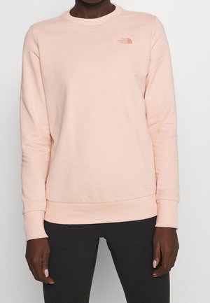 Person iført en lyserød langærmet sweatshirt med The North Face-logo på brystet og sorte bukser, stående mod en ensfarvet baggrund.