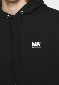 Sort hættetrøje lavet af bomuldsblanding, med et hvidt logo foran. Inkluderer metaløjer og snore til justering.