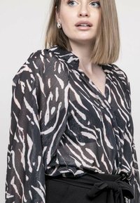 Blouse noire transparente ornée de rayures abstraites en zèbre blanches et rose clair. Avec un col et des manches longues. Portée sur une taille nouée noire.