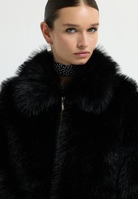 Czarna kurtka z sztucznego futra z szerokim kołnierzem i zapięciem na zamek. Posiada choker w kropki. Gładka tekstura z błyszczącym wykończeniem.