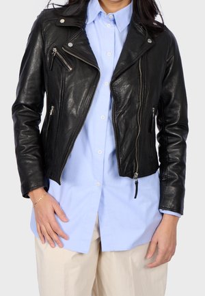 Mujer con chaqueta de cuero negra de motorista sobre una camisa azul clara abotonada y pantalones beige, manos relajadas a los lados.