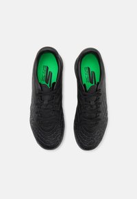 Schwarze Sportschuhe mit strukturierter Oberfläche, runder Zehenpartie und grünen Einlegesohlen mit Branding. Leichte Polsterung und Schnürverschluss.