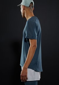 Lehká, texturovaná atletická košile v barvě teal s krátkými rukávy, bočními rozparky a minimalistickým designem. Doplněná světlou čepicí.