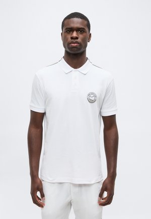 Noor mees seisab näoga otse, seljas valge polosärk, millel vasakul rinnal tennisklubi logo, taust on ühtlase valge värviga.