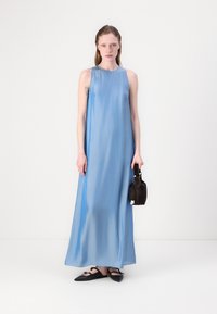 Long, sleeveless blue dress in a smooth, flowing fabric, paired with black flat shoes and a small, dark handbag. Minimalist design.