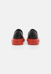Čierne kožené topánky s červenými gumovými podrážkami, ktoré majú elegantný dizajn a pohodlný slip-on štýl. Textúrované akcenty pri päte.